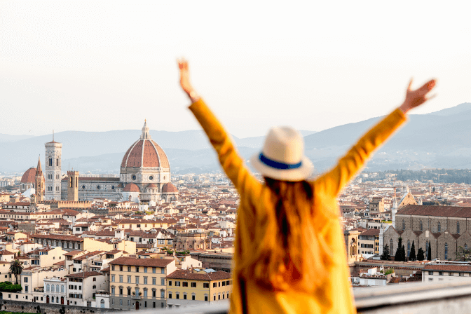 Woman with yellow jumper and hat standing infront of the Florence city skyline with hands up above her head.